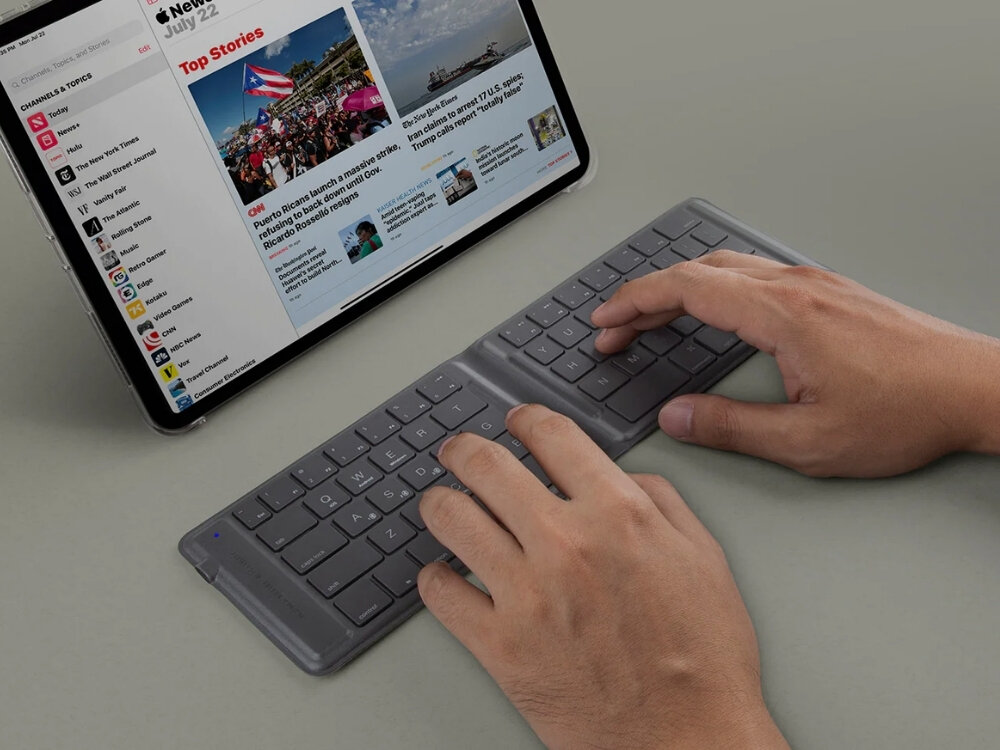 Osoba pisze na składanej klawiaturze podłączonej do tabletu, na którym wyświetlana jest strona z wiadomościami. Na ekranie widoczny jest tekst \'Top Stories\' oraz zdjęcia i artykuły.
