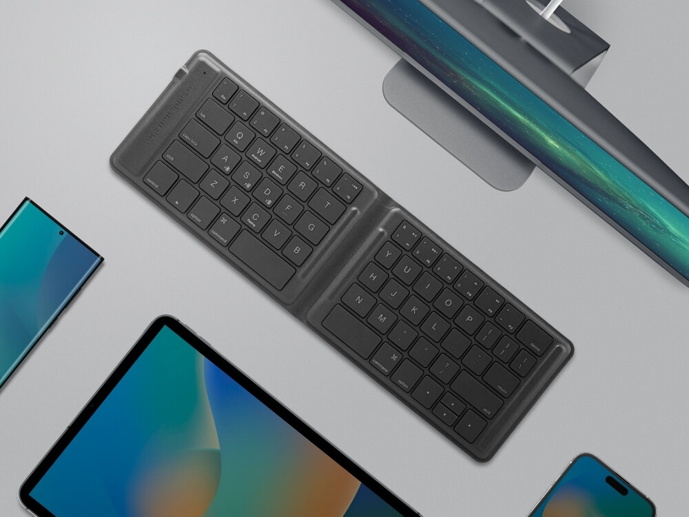 Na obrazie widoczna jest czarna klawiatura z układem QWERTY, umieszczona na biurku obok różnych urządzeń elektronicznych. Klawiatura ma składany design i znajduje się w otoczeniu monitorów i smartfonów.