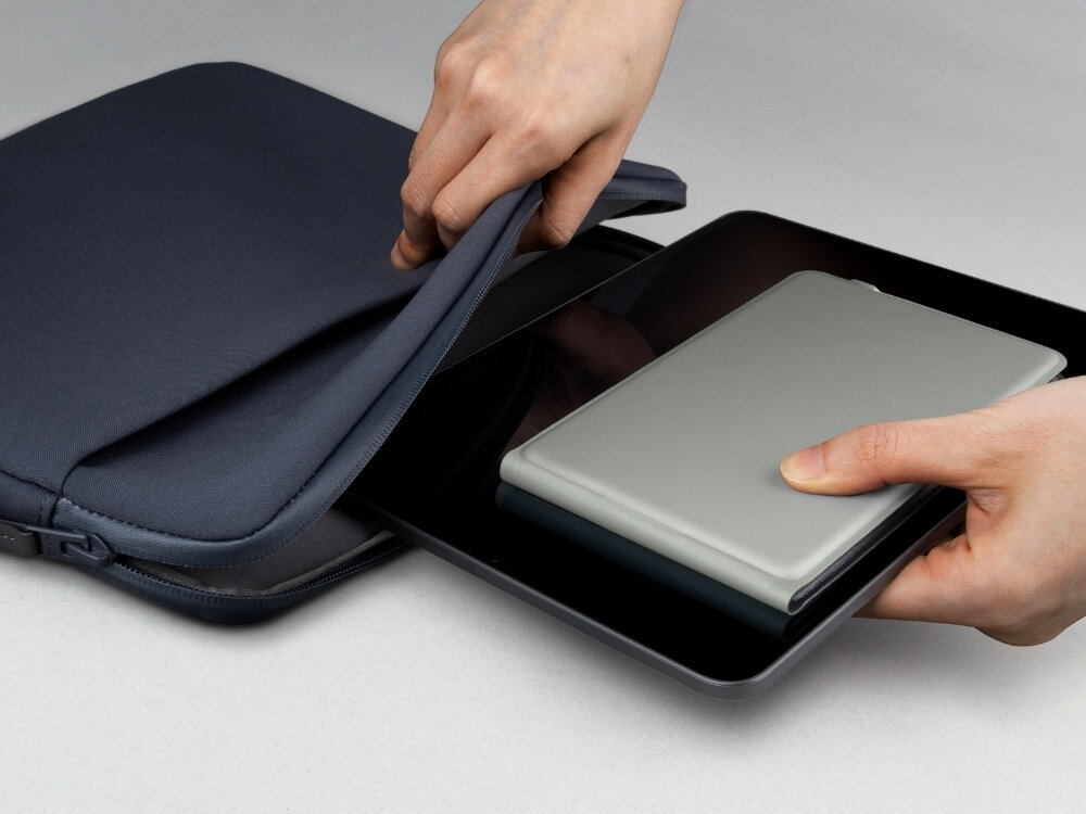 Osoba wkłada szary tablet do czarnej torby na laptopa. Torba jest otwarta, a tablet jest trzymany w dłoni.