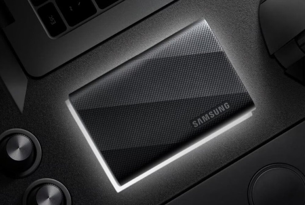 Na obrazie widoczny jest czarny dysk przenośny z teksturą, na którym znajduje się napis \'SAMSUNG\'. Dysk leży na ciemnej powierzchni obok innych urządzeń elektronicznych.