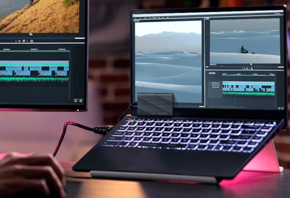 Na obrazie widać laptop z podłączonym zewnętrznym dyskiem SSD marki SAMSUNG, na ekranie wyświetlane są programy do edycji wideo. Obok znajduje się drugi monitor z podobnym oprogramowaniem.