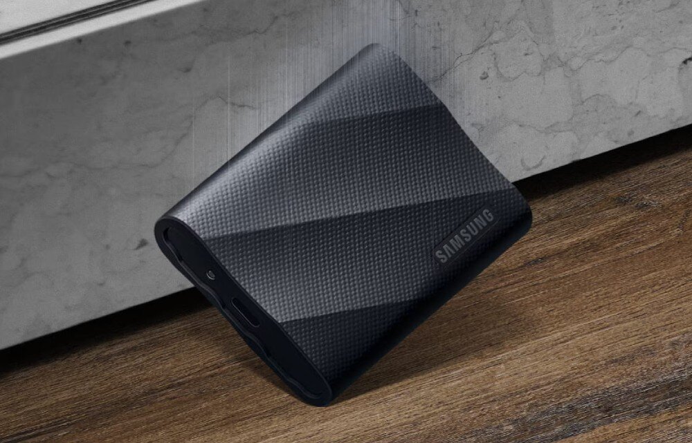 Czarny dysk przenośny z teksturą, z widocznym logo \'SAMSUNG\', leży na drewnianej powierzchni obok betonowej ściany.