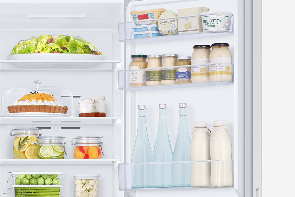 Wnętrze lodówki z widocznymi półkami wypełnionymi różnymi produktami spożywczymi, takimi jak sałata, ciasto, słoiki z przetworami, butelki z napojami oraz opakowania z serkiem kremowym i innymi produktami. Na drzwiach lodówki znajdują się półki z butelkami i słoikami.