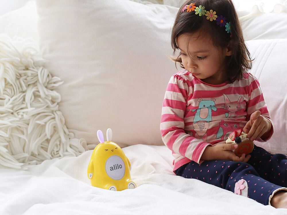 Zabawka edukacyjna ALILO Happy Bunny P1 Niebieski delikatne światło nocne, które pomaga w budowaniu poczucia bezpieczeństwa podczas 
					zasypiania i sprzyja spokojnemu wypoczynkowi, miękkie, świecące uszy króliczka, subtelne, zmieniające się światło, 7 kolorów
					dziewczynka siedząca na łóżku, obok niej produkt w wersji żółtej