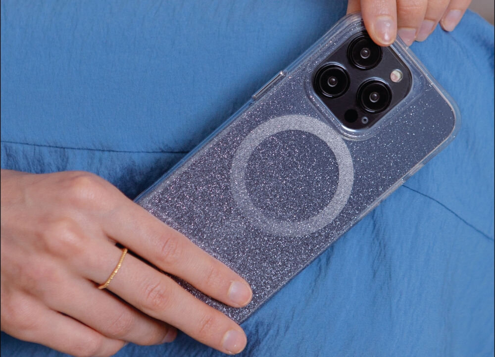 Osoba trzyma telefon w przezroczystym etui z brokatowym wykończeniem i widocznym okręgiem na plecach. Telefon ma trzy obiektywy aparatu i jest trzymany na tle niebieskiego materiału.