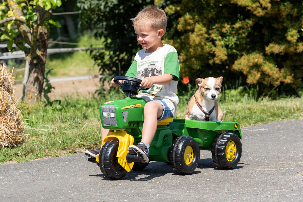 Przyczepa ROLLY TOYS Rolly Kipper John Deere droga park dziecko jedzie po drodze na traktorku ciągnie za sobą przyczepkę na przyczepce siedzi pies w tle drzewa zabawa rozwój atrakcja dla dziecka