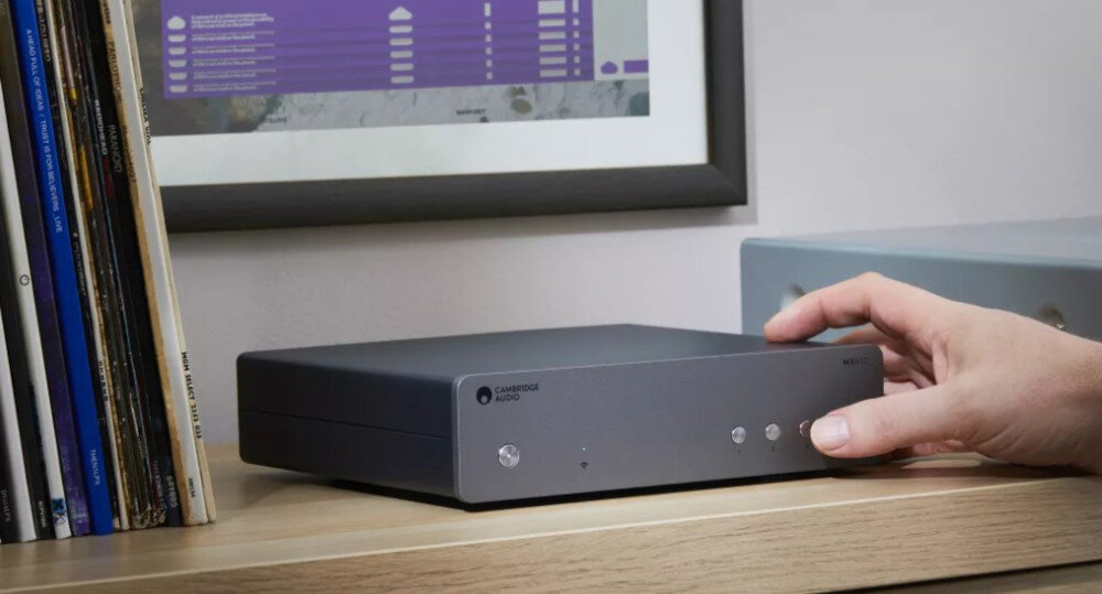 Odtwarzacz sieciowy CAMBRIDGE AUDIO MXN10 obsługa plików Hi-Res PCM DSD512 wysoka jakość dźwięku, odtwarzacz stojący obok czasopism, ręka trzymająca urządzenie