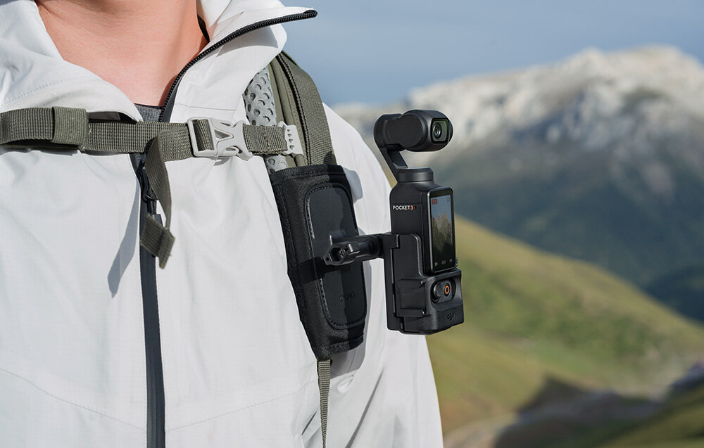 Osoba w białej kurtce ma zamocowaną na pasku plecaka kamerę DJI Osmo Pocket 3 z adapterem rozszerzającym. W tle widać górski krajobraz.