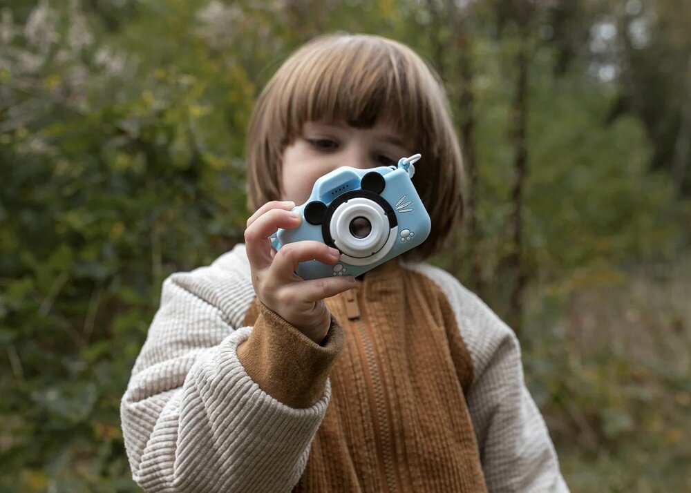 Dziecko trzyma niebieski aparat fotograficzny z motywem zwierzęcym, na tle zielonego otoczenia. Aparat ma duży obiektyw i ozdobne elementy przypominające uszy i wąsy.