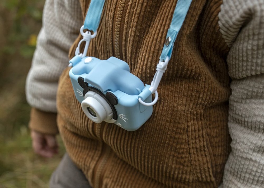 Na zdjęciu widać niebieski aparat dla dzieci zawieszony na szyi osoby, z białym obiektywem i ozdobami przypominającymi uszy oraz wąsy. Aparat jest przymocowany do niebieskiego paska.