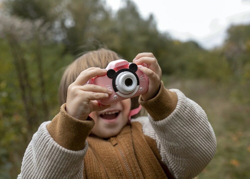 Dziecko trzyma różowy aparat fotograficzny z ozdobami przypominającymi uszy myszy. W tle widoczna jest rozmyta zieleń roślinności.