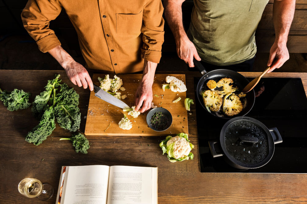 Nóż FISKARS Hard Edge 1051747 osoba kroi kalafior na desce druga osoba smazy warzywo na patelni Ostrze o imponującej  z wysokiej jakości japońskiej stali nierdzewnej precyzja efektywność trwałość dynamiczne cięcie z dużą siłą subtelna precyzja w codziennym gotowaniu przy wymagających technikach kulinarnych