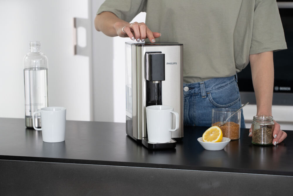 Osoba obsługuje dyspenser do wody marki PHILIPS, umieszczony na blacie obok kubka, butelki z wodą, szklanki z cukrem, cytryny i słoika z herbatą. Na urządzeniu widoczne jest logo PHILIPS.
