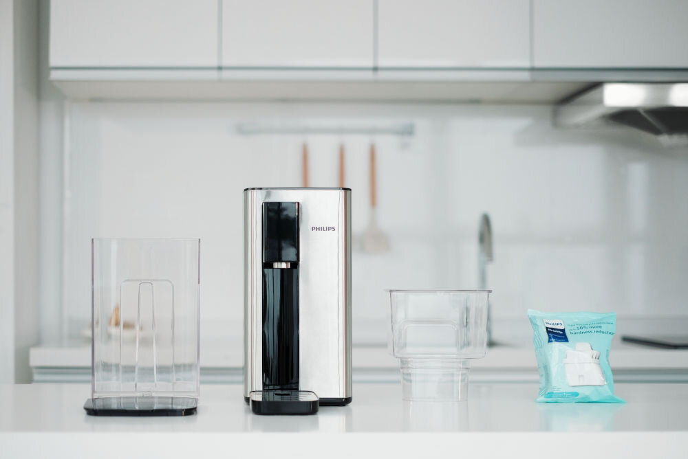 Na blacie kuchennym znajduje się dyspenser do wody marki PHILIPS, obok przezroczysty pojemnik i opakowanie z filtrem. W tle widoczna jest nowoczesna kuchnia z białymi szafkami.