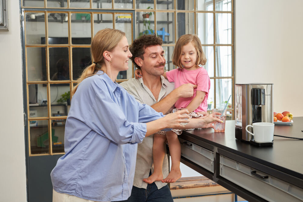 Na blacie kuchennym stoi dyspenser do wody obok kubka i szklanki. W tle widać osoby w kuchni, a na blacie leżą owoce.