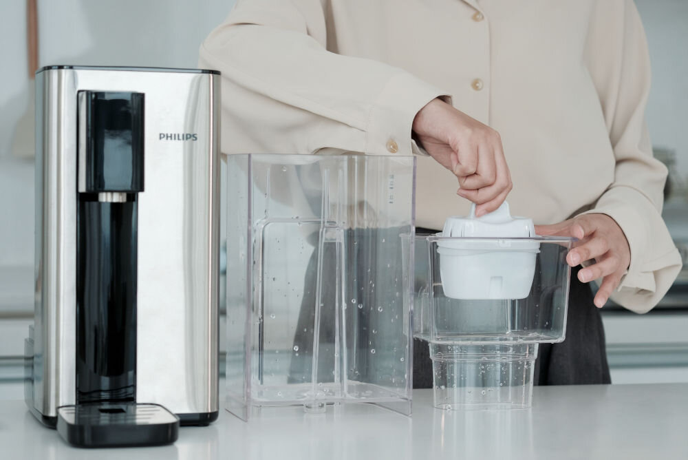 Osoba obsługuje przezroczysty pojemnik z filtrem w pobliżu dyspensera do wody marki PHILIPS. Dyspenser ma metaliczną obudowę i czarny panel przedni.