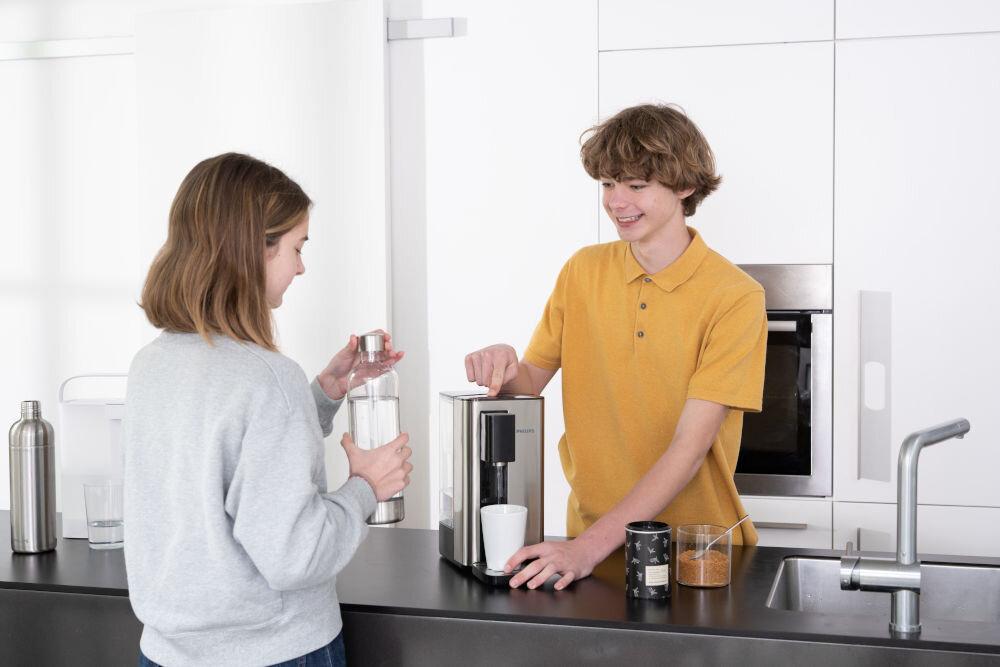 Dwie osoby stoją przy blacie kuchennym, na którym znajduje się dyspenser do wody z kubkiem pod wylewką. Obok dyspensera leżą dwa słoiki z przyprawami i szklanka z wodą.