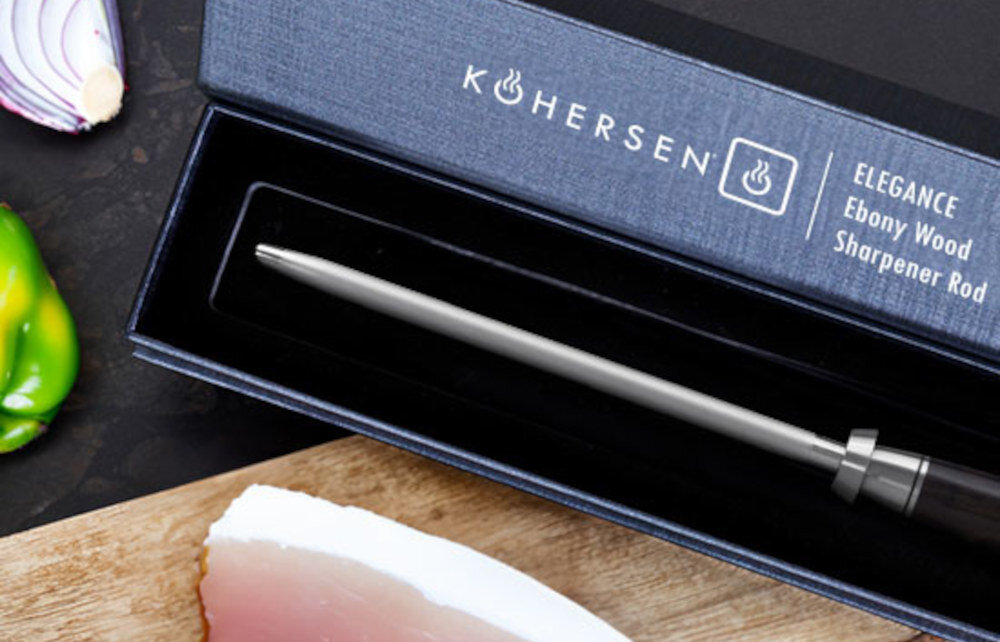 Ostrzałka KOHERSEN Elegance w eleganckim pudełku z napisem \'KOHERSEN ELEGANCE Ebony Wood Sharpener Rod\'. Obok pudełka znajdują się kawałki cebuli i zielonej papryki oraz fragment deski z jedzeniem.