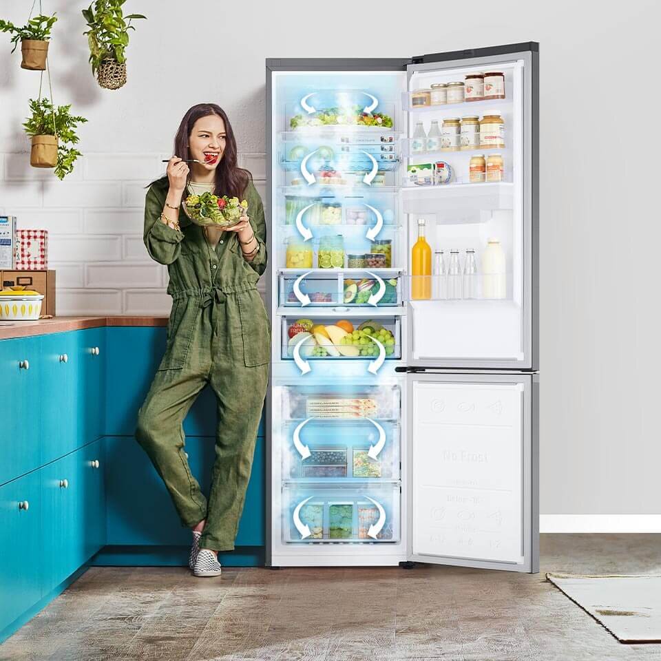 Kobieta w zielonym kombinezonie stoi obok otwartej lodówki, jedząc sałatkę. Wnętrze lodówki jest wypełnione różnorodnymi produktami spożywczymi, a na drzwiach widoczne są butelki i słoiki.