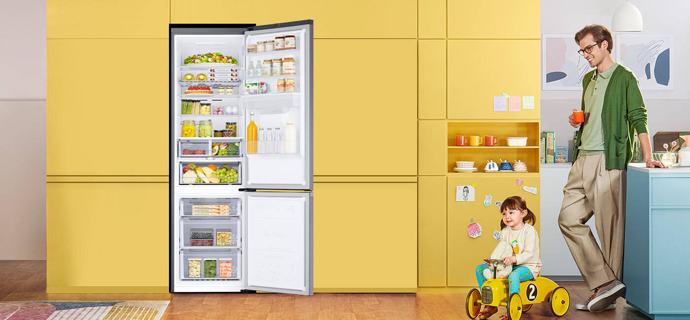 Otwarta lodówka wypełniona różnorodnymi produktami spożywczymi stoi na tle żółtej ściany. Obok znajduje się dziecko na żółtym rowerku oraz osoba dorosła z kubkiem w ręku.