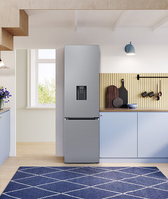 W kuchni znajduje się lodówka w kolorze szczotkowanej stali nierdzewnej z podajnikiem wody na drzwiach. Obok lodówki są niebieskie szafki kuchenne i dekoracyjne deski do krojenia na blacie.