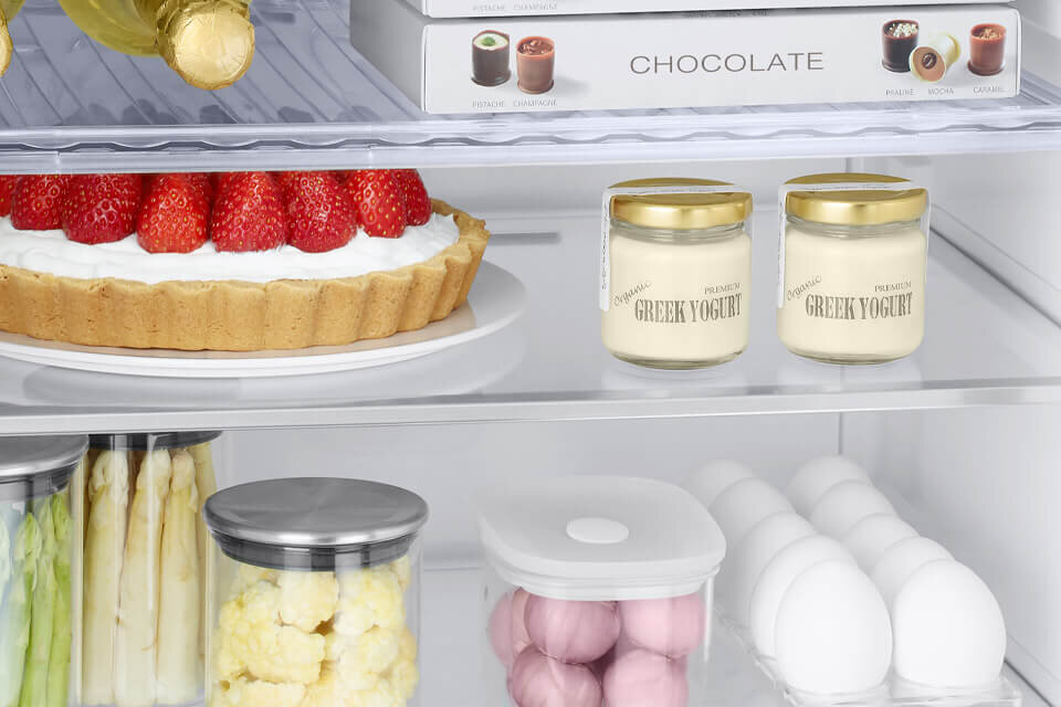 W lodówce znajdują się różne produkty spożywcze, w tym tarta z truskawkami, słoiki z jogurtem greckim oraz opakowanie czekoladek z napisem \'CHOCOLATE\'.