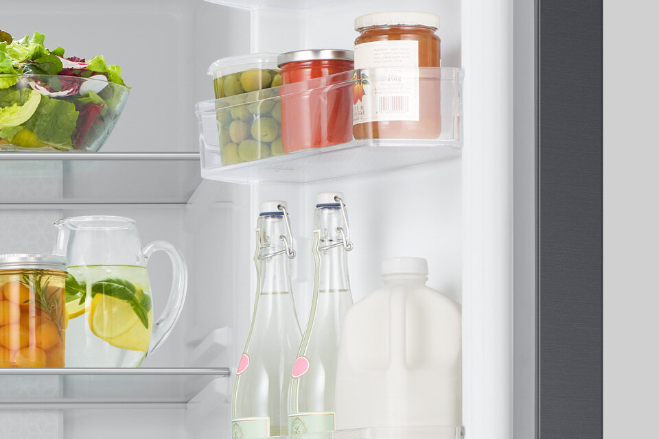 Wnętrze lodówki z widocznymi półkami, na których znajdują się słoiki z przetworami, butelki z napojami oraz dzbanek z wodą i cytryną. Na jednej z półek stoi miska z sałatą.