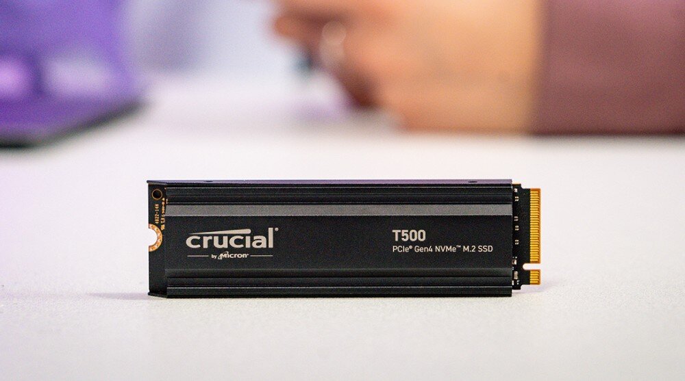Dysk CRUCIAL T500 Czarny dysk SSD stoi pionowo na jasnej powierzchni, w tle niewyraźna sylwetka osoby i laptop, pojemność, Microsoft DirectStorage