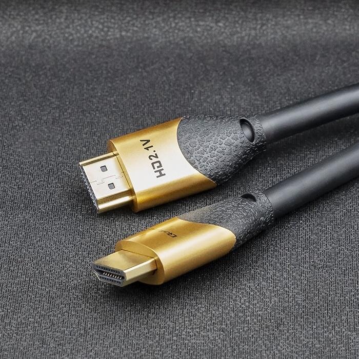 Na obrazie widoczny jest kabel HDMI z końcówkami w kolorze złotym i czarnym, z napisem \'HD2.1V\' na jednej z nich. Kabel leży na szarej powierzchni.