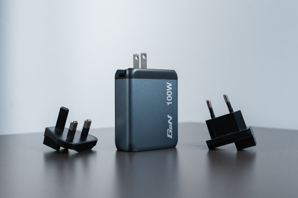 Na obrazie widoczna jest szara ładowarka z napisem \'GaN 100W\' oraz dwa adaptery wtyczek. Ładowarka i adaptery znajdują się na gładkiej powierzchni.