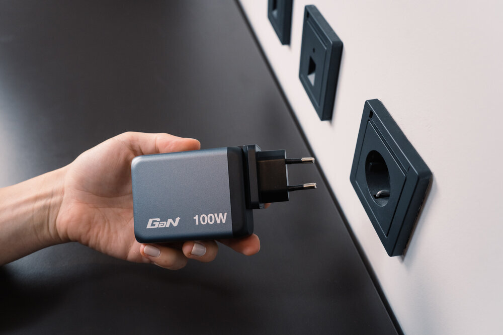 Osoba trzyma szarą ładowarkę z napisem \'GaN 100W\' przed gniazdkiem elektrycznym w ścianie.
