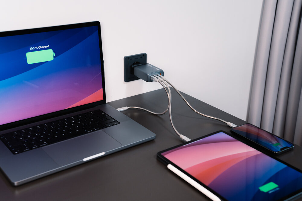 Na biurku znajduje się laptop z wyświetlonym napisem \'100% Charged\', tablet i smartfon, wszystkie podłączone do szarej ładowarki w gniazdku ściennym. Ładowarka ma kilka kabli podłączonych do różnych urządzeń.