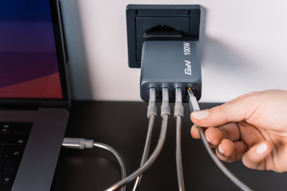 Szara ładowarka z napisem \'GaN 100W\' podłączona do gniazdka, z kilkoma kablami wpiętymi do portów. Osoba trzyma jeden z kabli, a obok znajduje się laptop.