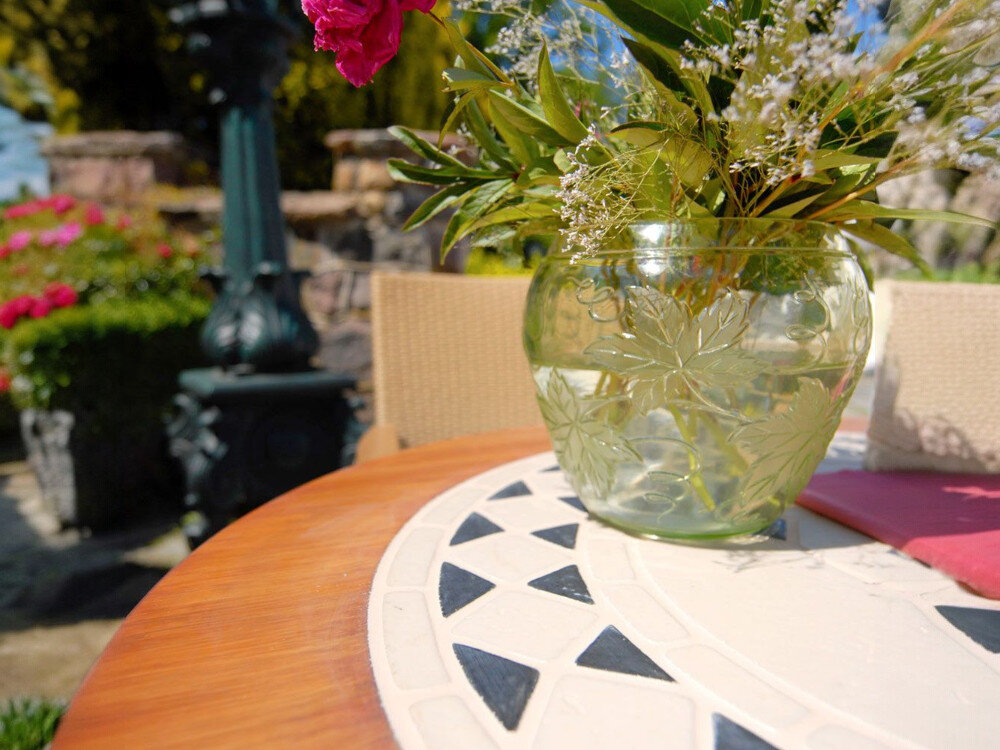 Stół ogrodowy PATIO Como 100 cm Brązowo-beżowy, UV, temperatura, ochrona, taras, dzbanek z roślinami stoi na stole, w tle roślinność