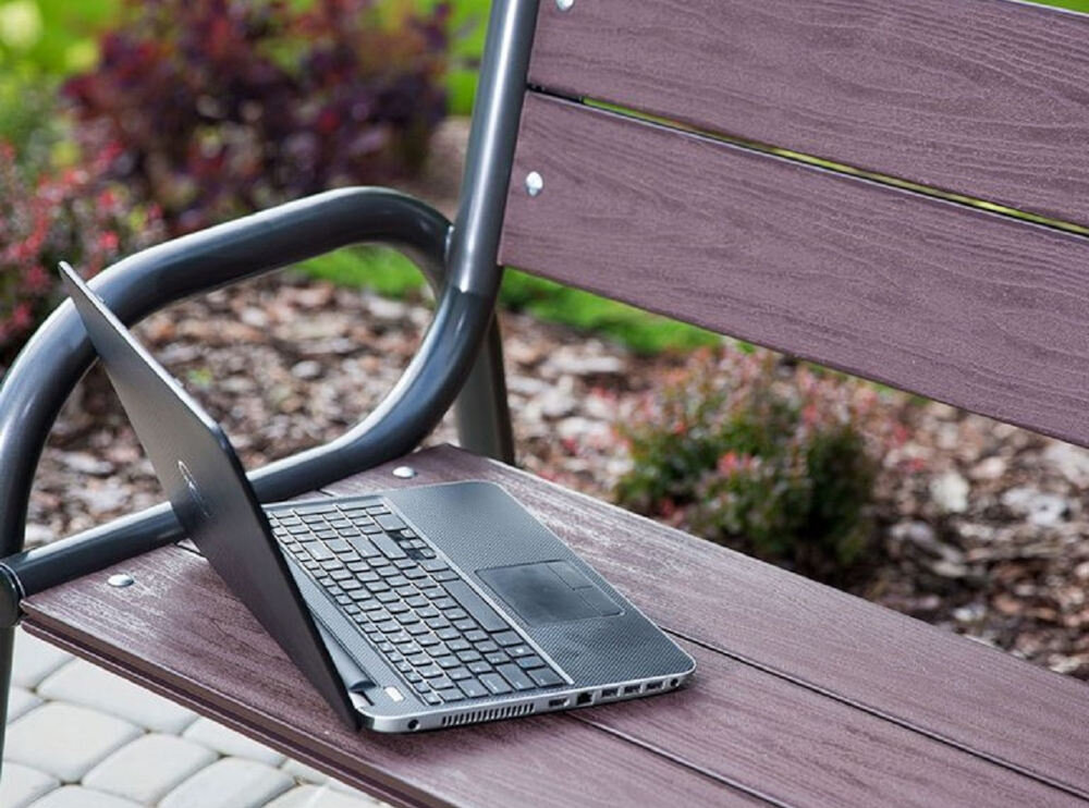 Na drewnianej ławce ogrodowej znajduje się otwarty laptop. Ławka ma metalowe podłokietniki i jest umieszczona na tle roślinności.