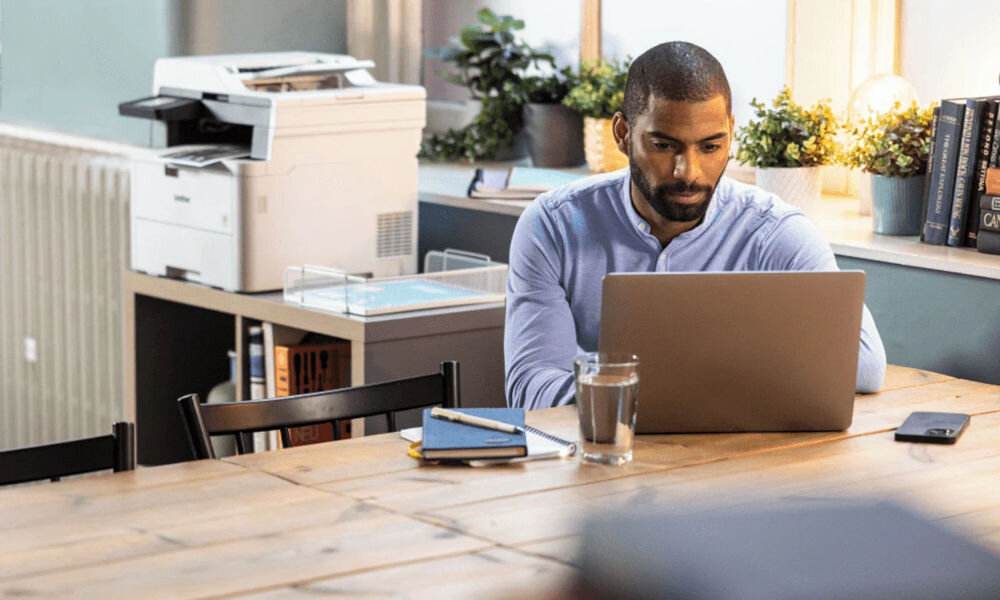 Urządzenie wielofunkcyjne BROTHER MFC-L3740CDW Druk w kolorze, Automatyczny druk dwustronny, Wi-Fi prezentacja urządzenia w biurze na półce przed mężczyzna siedzący przy stole z laptopem