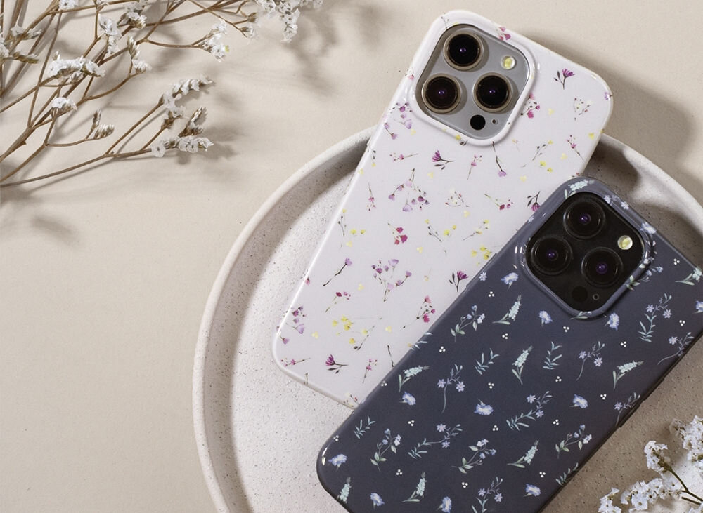 Na obrazie znajdują się dwa etui na telefon z motywem kwiatowym, jedno w kolorze beżowym, drugie ciemnym. Etui leżą na okrągłej tacy, a w tle widoczne są suche gałązki roślin.