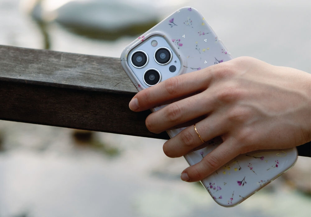 Osoba trzyma telefon w beżowym etui z delikatnym wzorem kwiatowym. W tle widoczna jest rozmyta sceneria z elementami natury.