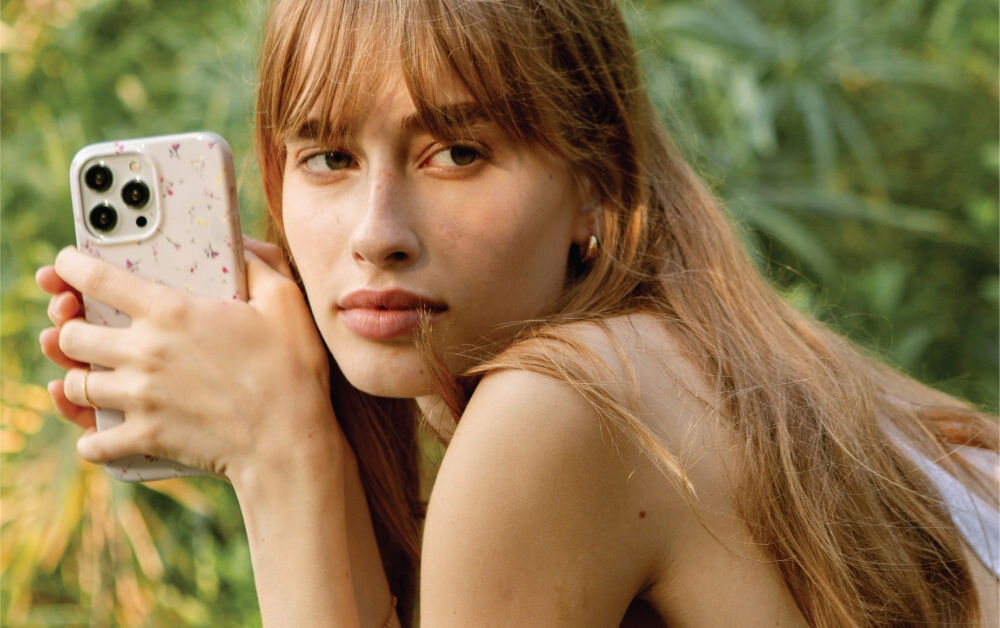 Osoba trzyma telefon w etui z delikatnym wzorem kwiatowym. W tle widoczna jest zielona roślinność.