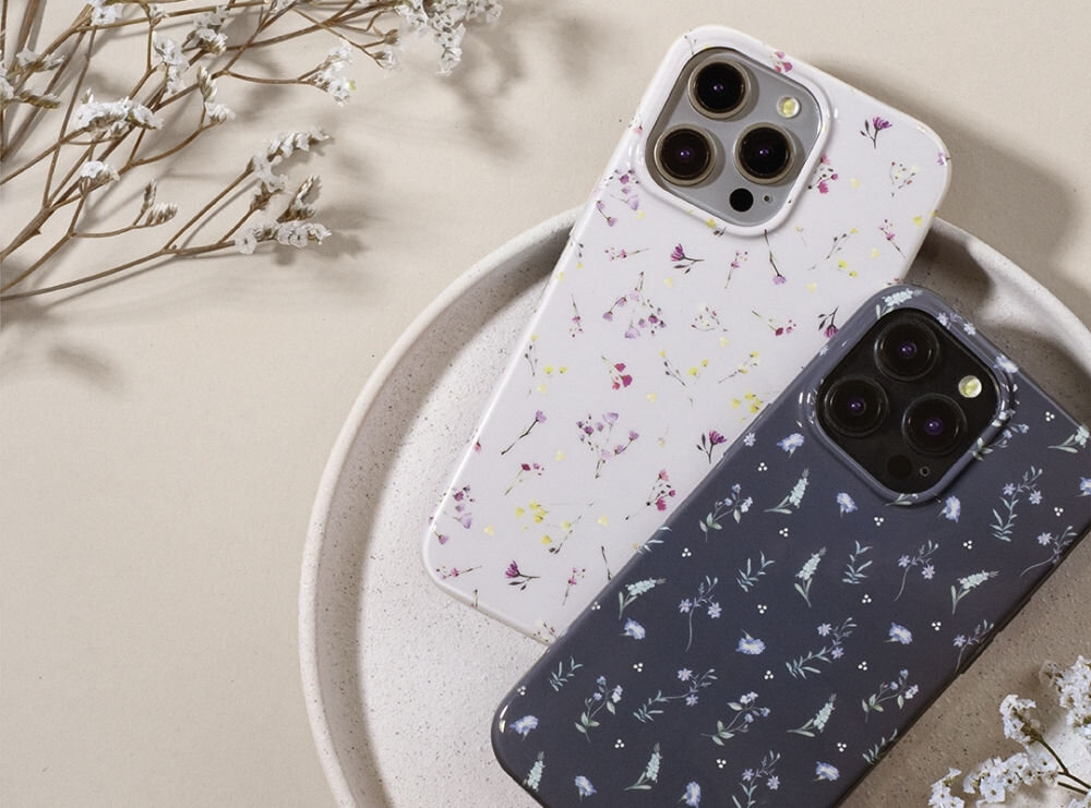 Na obrazie widoczne są dwa etui na telefon z motywem kwiatowym, jedno w kolorze białym, drugie granatowym. Etui leżą na okrągłej tacy, a w tle znajdują się suszone kwiaty.