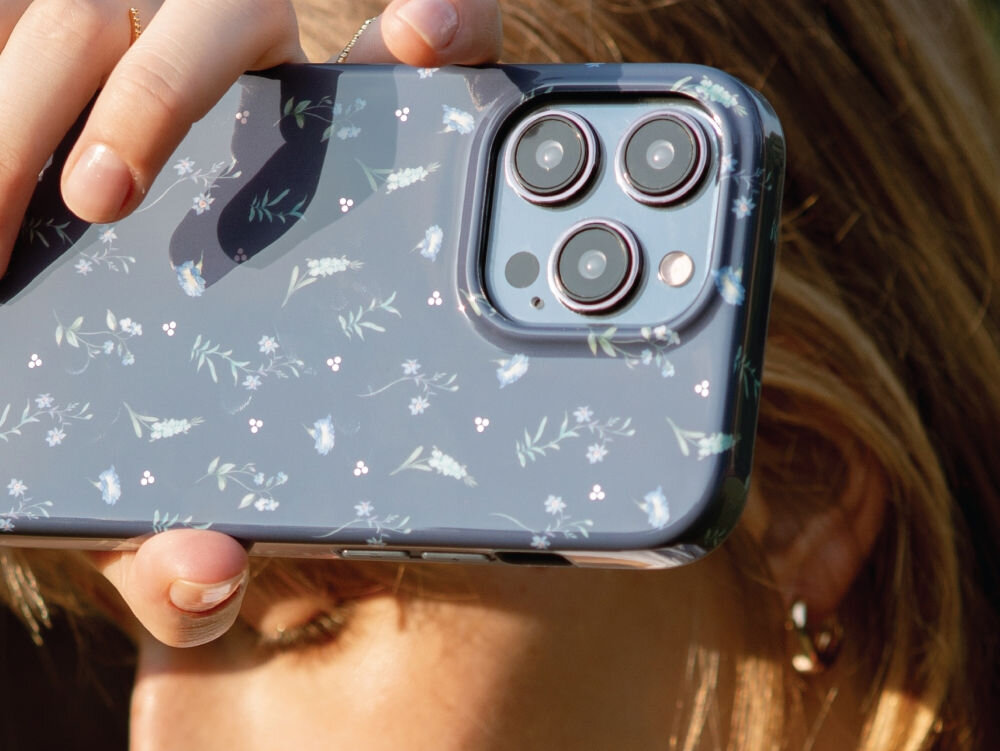 Osoba trzyma telefon z granatowym etui ozdobionym delikatnym wzorem kwiatowym. Widoczne są trzy obiektywy aparatu telefonu.