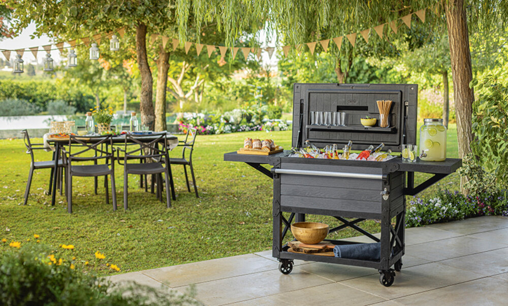 Barek ogrodowy KETER PATIO COOLER & BEVERAGE 255187 barek z otwartą klapą wnętrze wypełnione lodem i butelkami w tle stół ogrodowy drzewa i krzewy sprzęt na każdą okazję wygoda chłodzenie napojów stolik spotkanie z przyjaciółmi