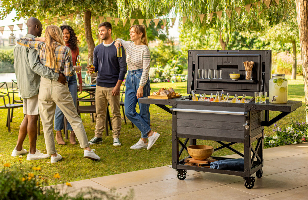 Barek ogrodowy KETER PATIO COOLER & BEVERAGE 255187 barek z otwartą klapą wnętrze wypełnione lodem i butelkami w tle kilka osob rozmawiających ze sobą ogród organizacja spotkań na świeżym powietrzu funkcja podręcznego stolika wygodny barek z pojemnikiem do chłodzenia napojów