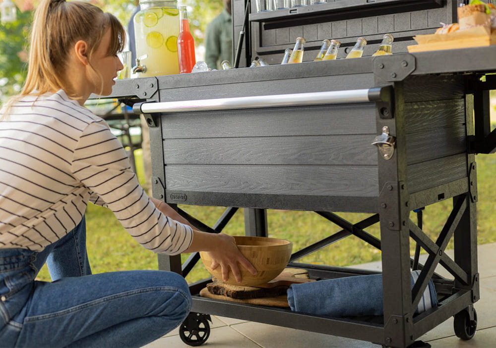 Barek ogrodowy KETER PATIO COOLER & BEVERAGE 255187 osoba kładzie miskę na dolnej półce barek wypłniony butekami i przekąskami dwie boczne półki udźwig do 15 kg każda dolna półka udźwig do 25 kg niewielka półeczka na wewnętrznej części klapy udźwig do 5 kg