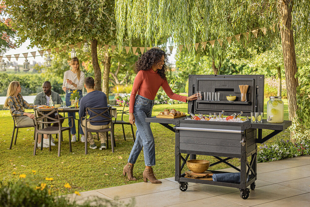 Barek ogrodowy KETER PATIO COOLER & BEVERAGE 255187 barek z otwartą klapą wnętrze wypełnione lodem i butelkami osoba podchodzi do barku w tle inne osoby przy stole i drzewa z trwałego tworzywa sztucznego odporne na wilgoć promieniowanie UV i zmienne temperatury nie odkształca się nie blaknie pod wpływem słońca gładka powierzchnia barku ułatwia jego czyszczenie przetrzeć ją wilgotną ściereczką lub spłukać wodą