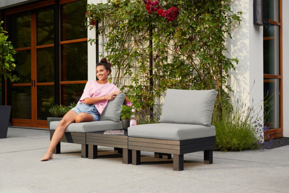 Zestaw mebli ogrodowych KETER 1 Elements 257366 Grafitowy zestaw na tarasie siedziska oddzielone stolikiem na jednym siedzi kobieta w tle dom i rośliny odporność na zabrudzenia łatwe do wyczyszczenia oszczędność czasu i wysiłku oszczędność ekologia