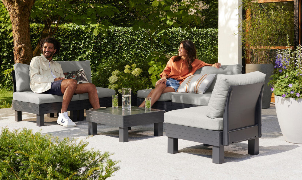 Zestaw mebli ogrodowych KETER 1 Elements 257366 Grafitowy taras dwie osoby każda siedzi na osobnym module w tle żywopłot dom i rośliny grafitowy stelaż jasnoszare poduszki elegancka neutralna kompozycja kolorystyczna dopasujesz do różnych stylów aranżacyjnych zestaw nie jest składany odporność na pogodę na zewnątrz przez cały sezon nie wymaga konserwacji ani zabezpieczenia