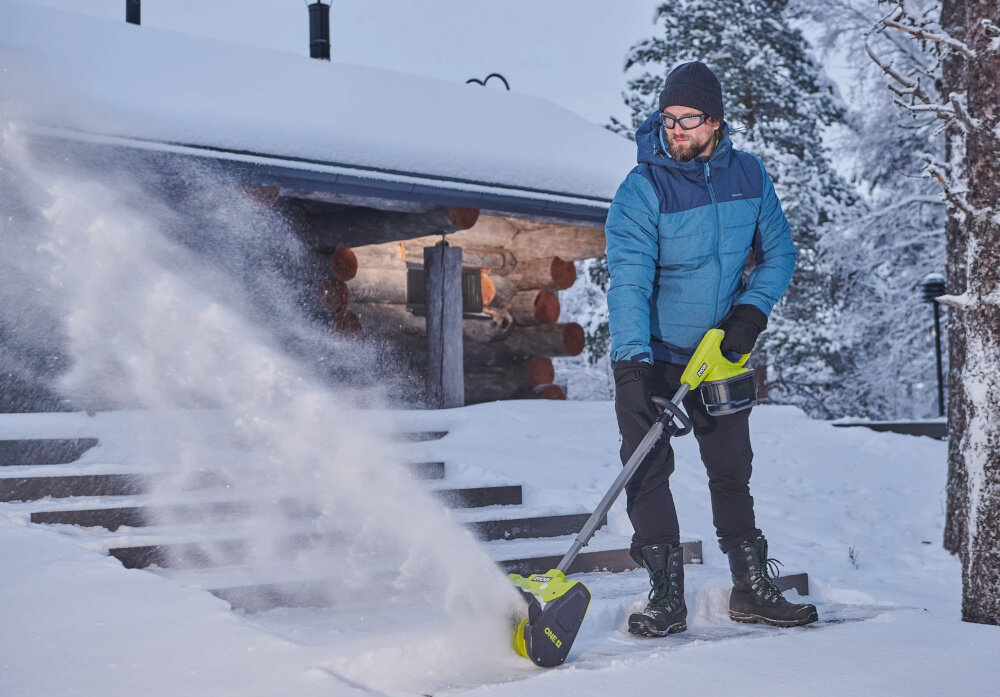 Odśnieżarka akumulatorowa RYOBI RY18ST25A-140 podwórko dom osoba odśnieża schody przed domem w tle drewniana ściana domu ośnieżone drzewa odśnieżanie bez wysiłku usuwanie śniegu skuteczność