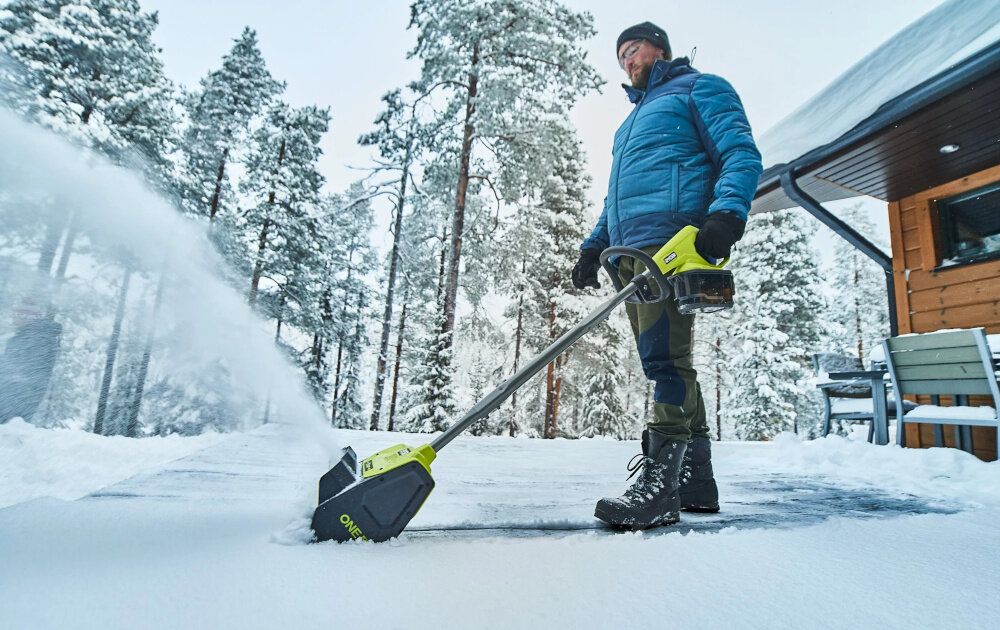 Odśnieżarka akumulatorowa RYOBI RY18ST25A-140 podwórko dom osoba odśnieża taras przed domem w tle drewniana ściana domu ośnieżone drzewa obudowa akumulatora o stopniu ochrony IPX4 zabezpiecza go przed wilgocią oraz zachlapaniem bezpieczeństwo użytkowania i stabilność zasilania