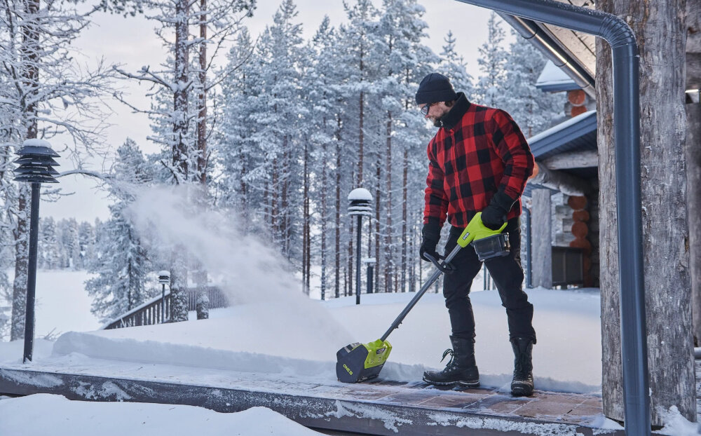 Odśnieżarka akumulatorowa RYOBI RY18ST25A-140 podwórko dom osoba odśnieża taras przed domem w tle drewniana ściana domu ośnieżone drzewa do pracy z lekkim i świeżym śniegiem o głębokości do 15 cm system pracy ogranicza konieczność używania siły fizycznej